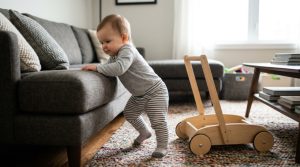 Smiling baby taking first steps on living room carpet toward parents hands.