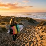 A surfer walks down a sandy path toward the ocean holding a surfboard.