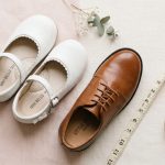 Childrens elegant white and brown wedding shoes next to a soft measuring tape.