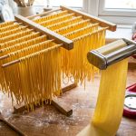 Hands guiding fresh dough through a KitchenAid pasta roller attachment.